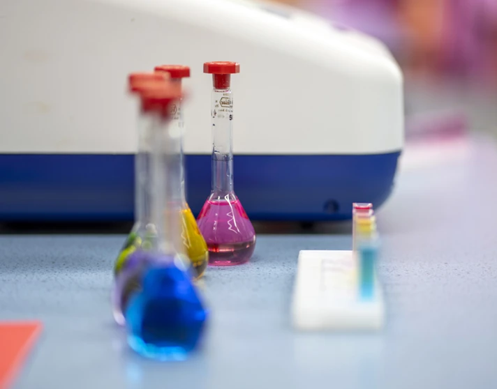 Three glass flasks filled with bright blue, yellow, and pink liquids sit on a lab bench, with a row of colourful test tubes and a science machine in the background—ready for a splash of science fun and discovery. Three glass flasks filled with bright blue, yellow, and pink liquids sit on a lab bench, with a row of colourful test tubes and a science machine in the background—ready for a splash of science fun and discovery.